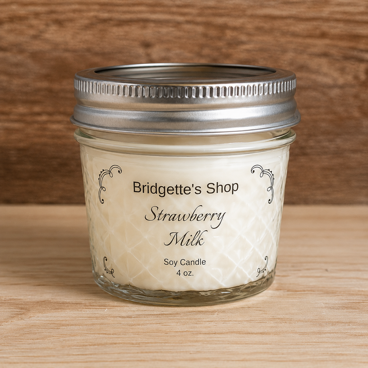 A 4 oz glass jar candle with a silver metal screw-top lid, labeled 'Bridgette's Shop Strawberry Milk Soy Candle'. The candle is set on a light wood surface with a darker wood background.