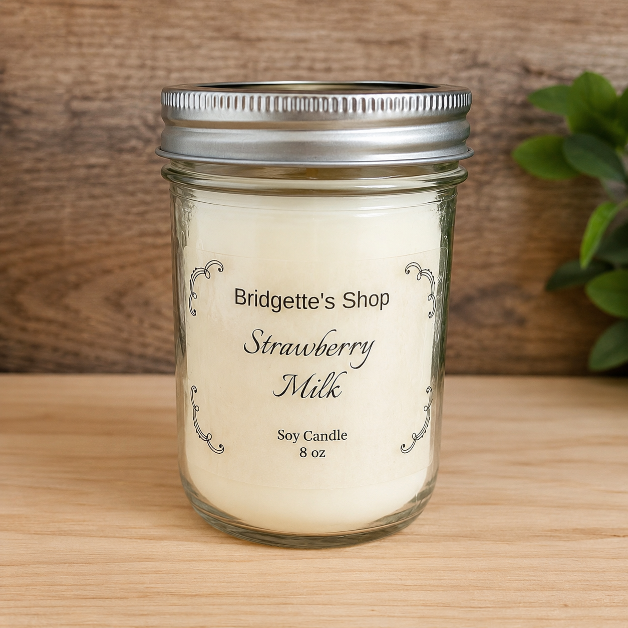 A 8 oz soy candle in a clear glass mason jar with a silver metal lid, labeled 'Bridgette's Shop Strawberry Milk'. The candle is set on a light wood surface with a rustic wooden background and a green plant leaf visible on the right.